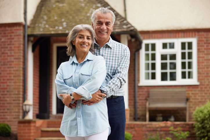 Portrait Of Mature Couple Standing In Garden In Front Of Dream Home In Countryside