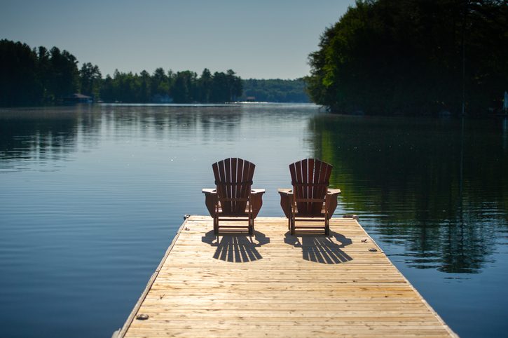Two Adirondack chairs sitting on a wooden pier facing the calm water of a lake in Muskoka, Ontario Canada. A cottage nestled between trees is visible in background.