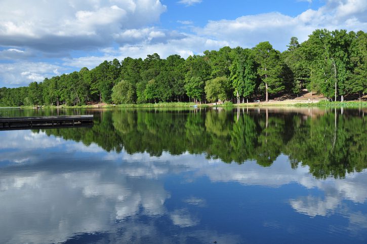 Peaceful scenic lake in East Texas.  The water reflects theReflects the blue sky and clouds.  The shoreline is full of green pine and oak trees.